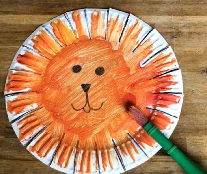 This easy art project for kids shows a paper plate that is painted orange with simple eyes, mouth, and nose painted on in black. The edges of the plate have been cut with slits to look like a mane.