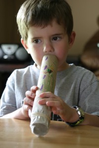 A little boy is seen blowing into a flute that is made from paper rolls.