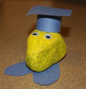 A rock is painted yellow and has a construction paper graduation cap and feet. It also has googly eyes.