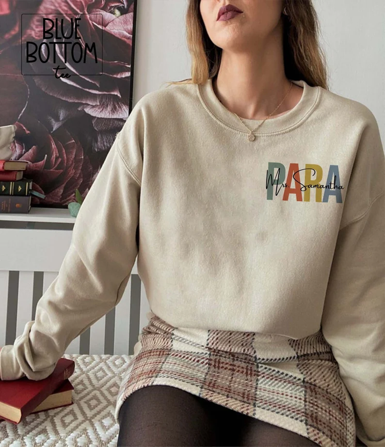 A woman is shown from the neck down. She is wearing an off white crewneck sweatshirt that says PARA and her name over it. (gifts for paraprofessionals)