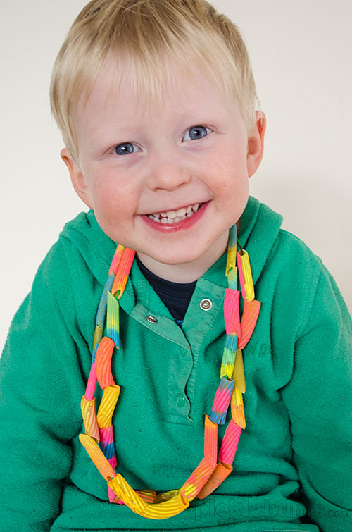 A small boy is wearing a necklace made from painted pasta and string.