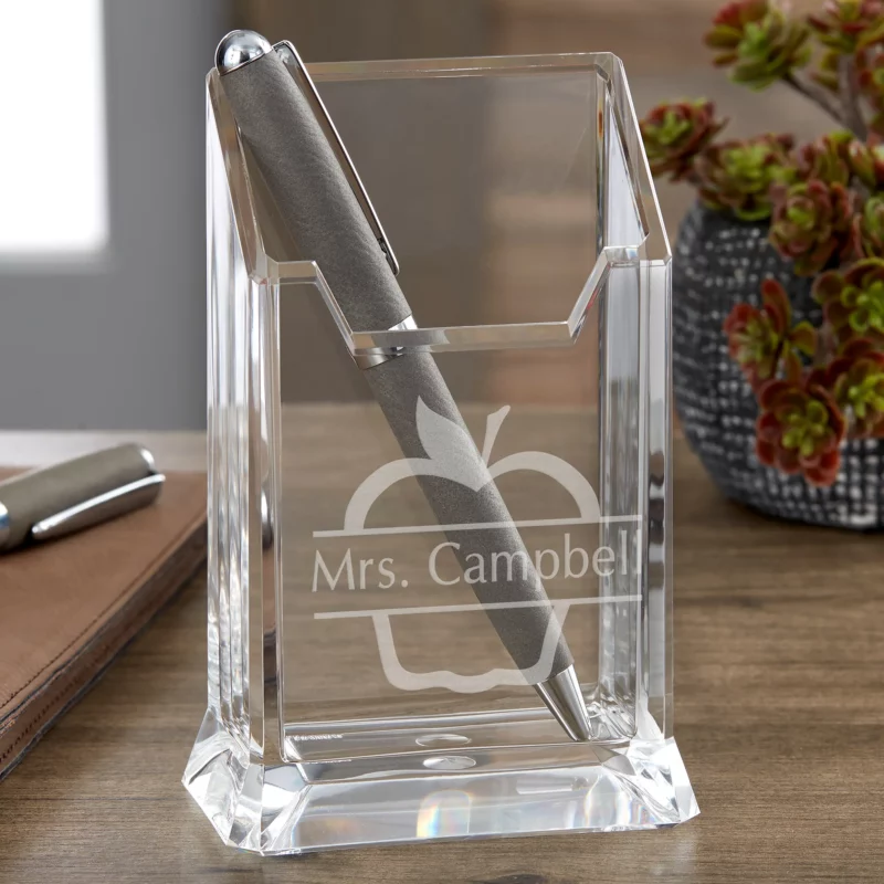 A clear glass pencil and pen holder has an apple silhouette and the name Mrs. Campbell