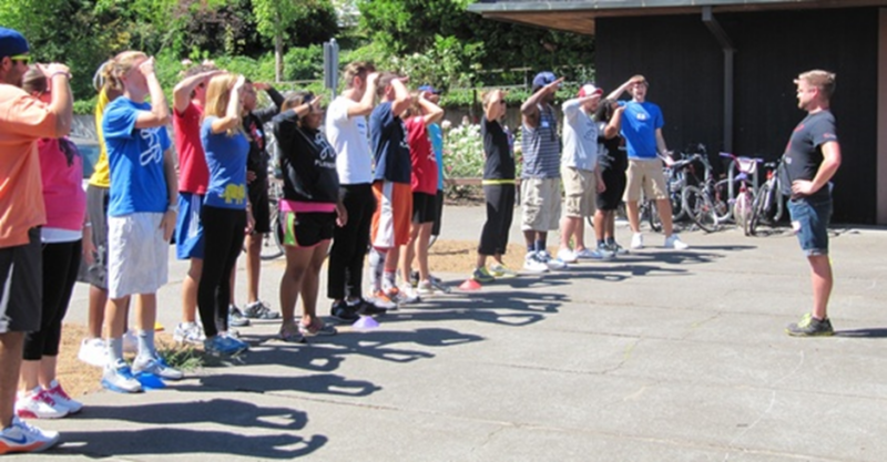 Several people stand facing an instructor outside.