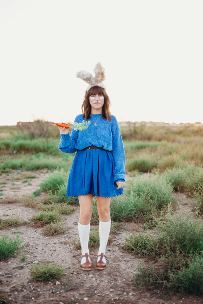 A woman is shown wearing bunny ears, holding a carrot, with bunny face paint and dressed in blue.