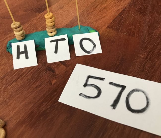 Uncooked spaghetti strands stuck upright into playdough and labeled H, T, and O with Cheerios stacked on each next to card reading 570