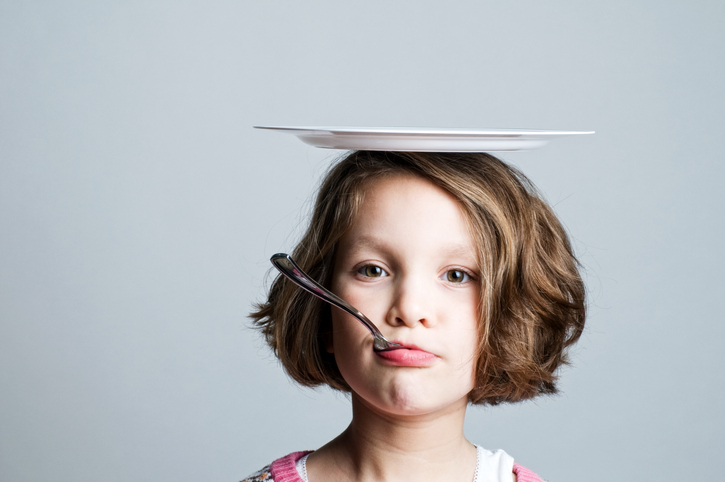 A young girl is shown from the neck up with a plate on her head and a spoon in her mouth.