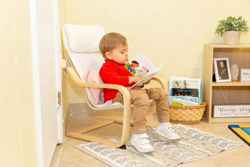 A small boy reads in a child sized white armchair.
