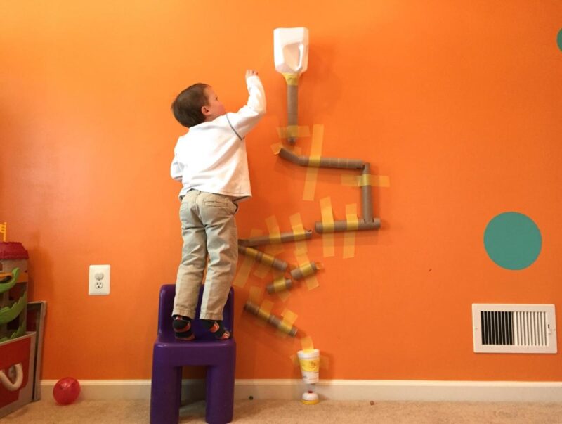 A boy standing on a chair reaching toward an orange wall with a pom pom drop attached