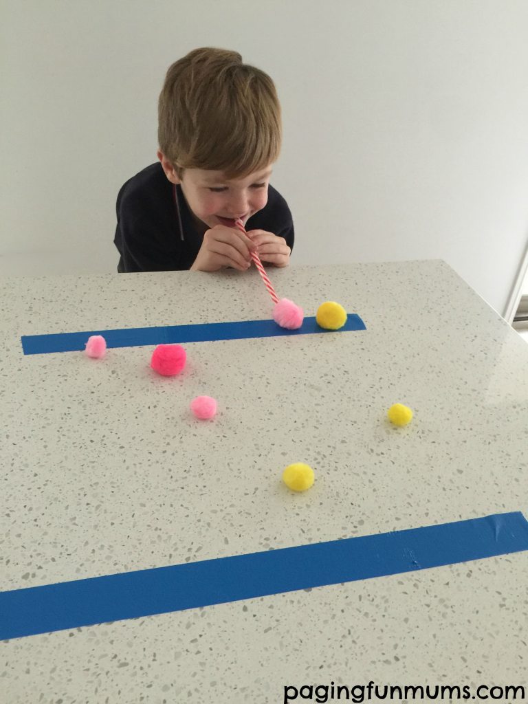 Minute to win it games include this one where a little boy blows yellow and pink pom poms across a table by blowing into a straw.
