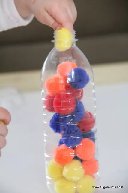 An empty water bottle is filled with pom poms and a little hand is shown placing another into the bottle (fine motor activities)