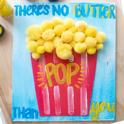 The best father's day crafts for kids should be homemade like this "There's no butter pop than you" painted on canvas to look like popcorn. Yellow pom poms have been added to look like popcorn.