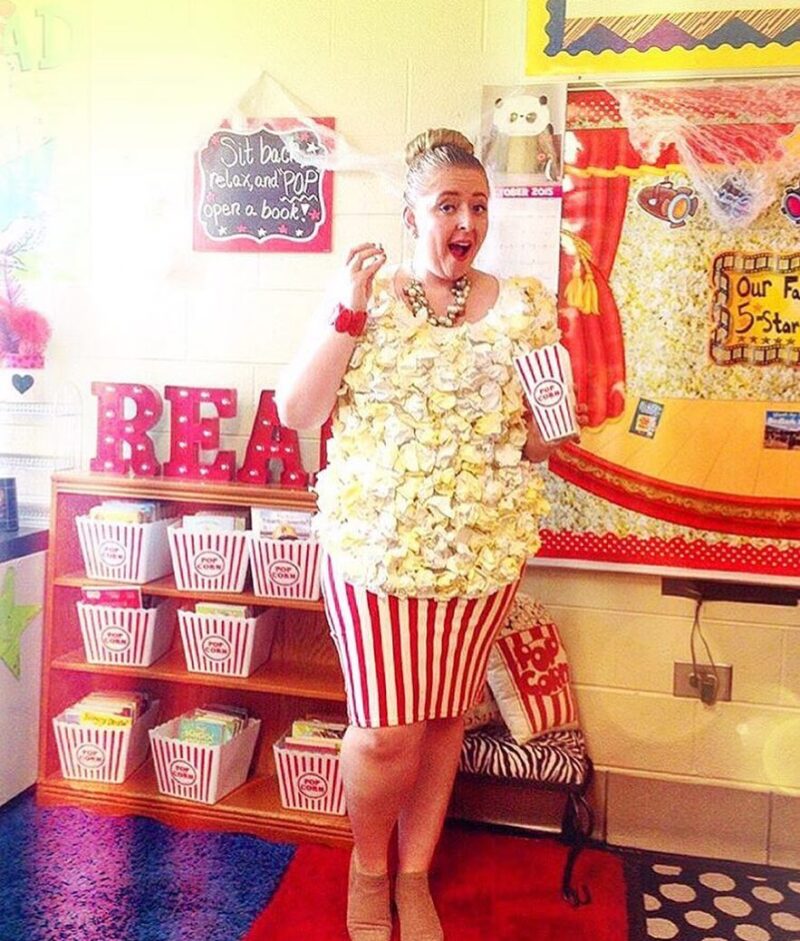A teacher is dressed as popcorn and is standing in front of a shelf of buckets of popcorn.