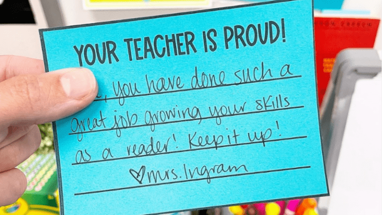 A hand holding a 'Your Teacher is Proud!' note on blue paper