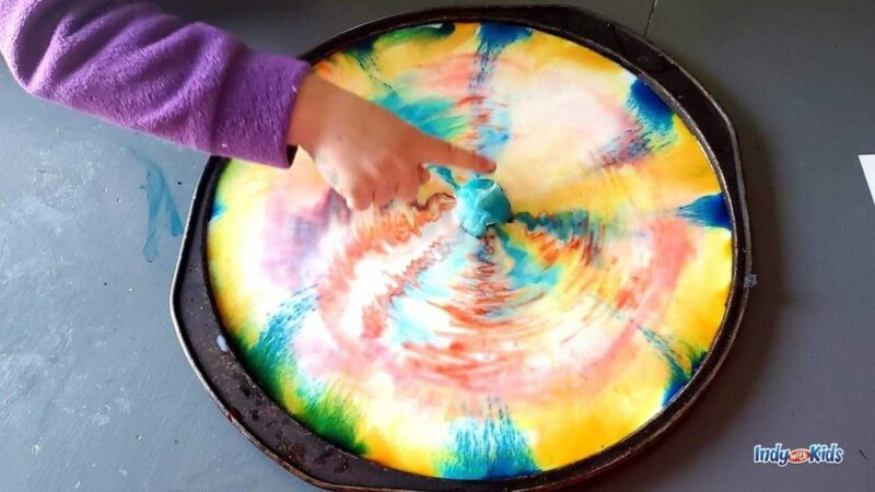 child's finger pointing to a rainbow swirl experiment, as an example of St. Patrick's Day activities 