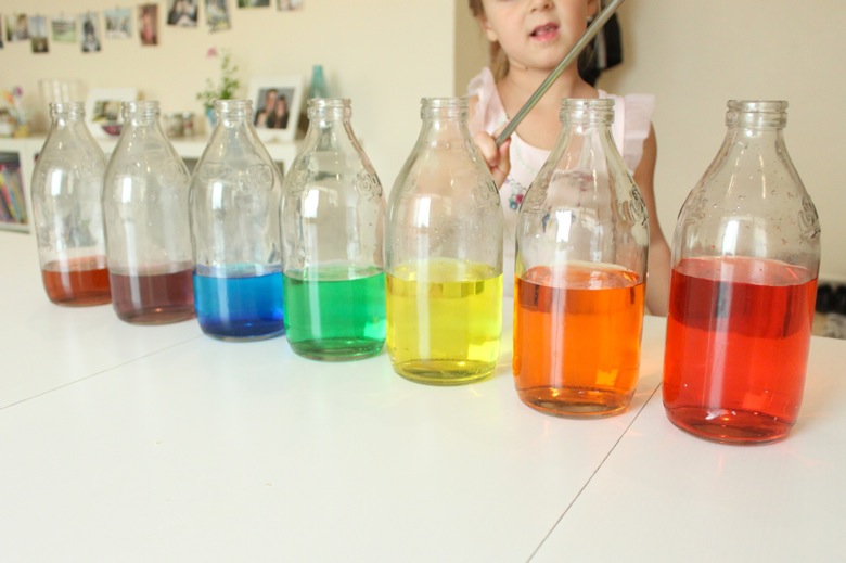 child standing behind row of glass jars with colored water in them 