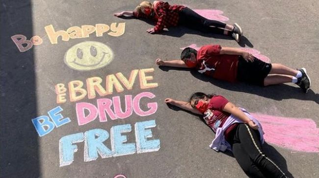 Three students lying by a chalk drawing saying Be happy, be brave, be drug-free (Red Ribbon Week Ideas)