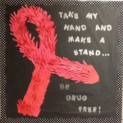 A large 3-D red ribbon made up of cut out red hands is shown on a black background with white text that reads "take my hand and make a stand, be drug free"