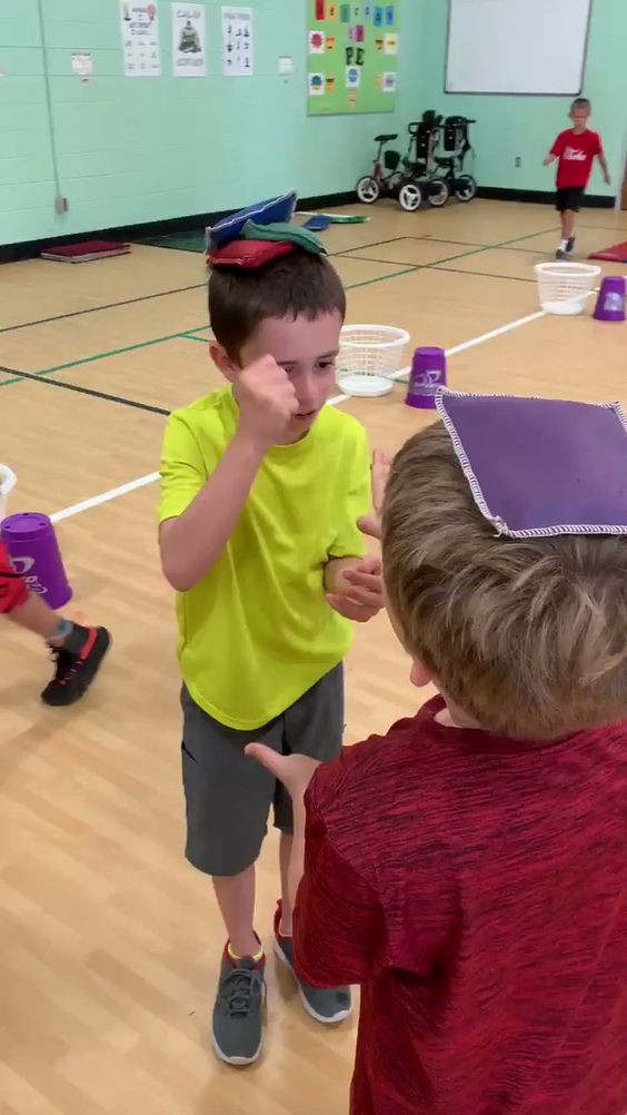 Two children stand playing rock, paper, scissors, with bean bags on their heads (elementary PE games)