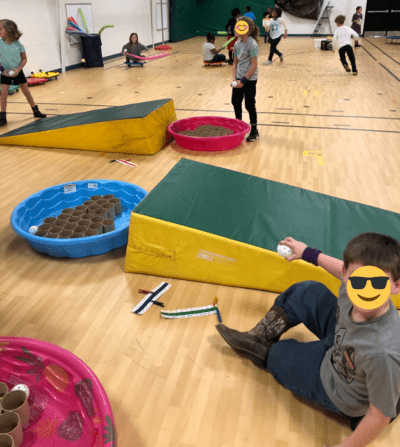 Wedge mats are laid out in front of kiddie swimming pools which are filled with industrial sized paper towel rolls. Children are scattered around holding whiffle balls.
