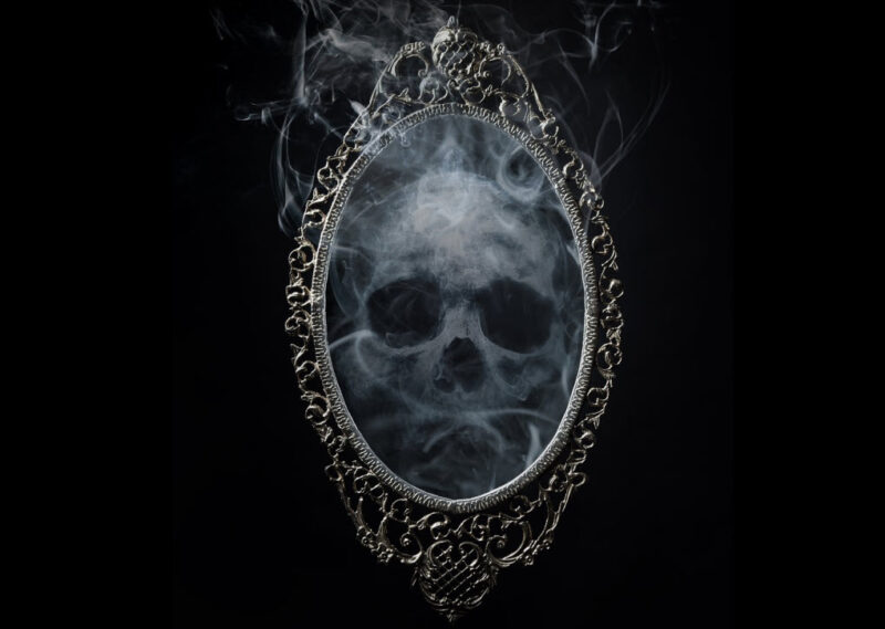 A spooky skull reflected in an ornate oval mirror against a black background