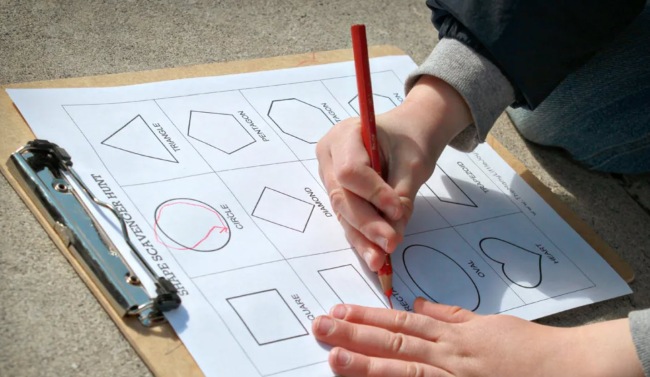 Child writing on a shapes scavenger hunt sheet attached to a clipboard