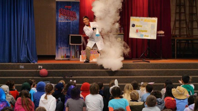 a man at a school assembly doing a science experiment that makes a cloud of steam for teaching 4th grade 
