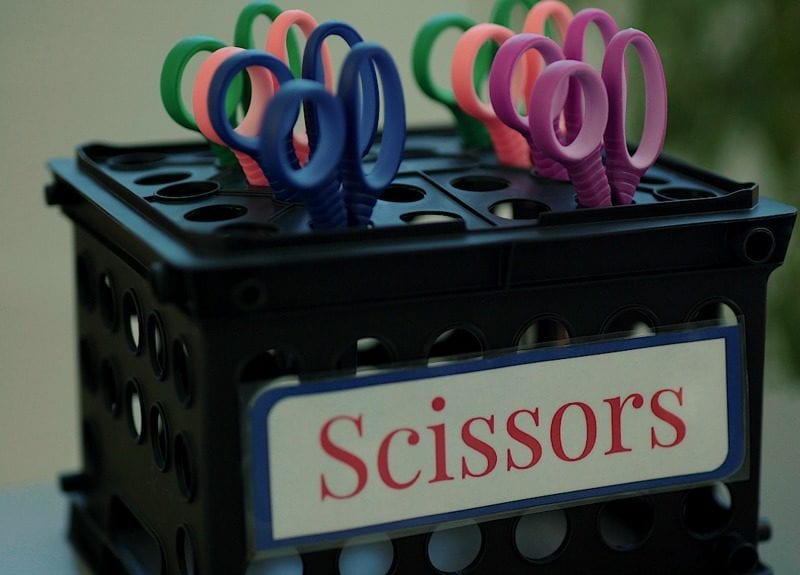 Black storage crate turned upside-down with scissors inserted into the holes, labeled Scissors