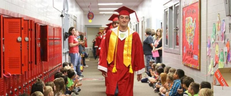 Students wearing graduation caps and gowns walk along the hall of their elementary school as teachers and students watch and applaud