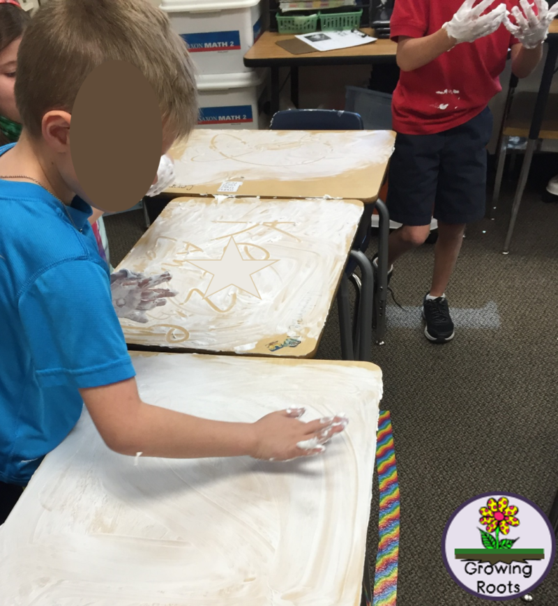 A student in a blue shirt writes in shaving cream on his desk with his finger