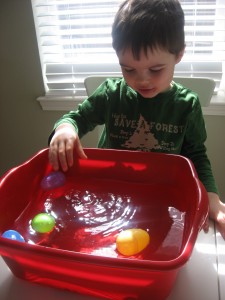 A little boy stands in front of a red tub filled with water. There are various plastic Easter eggs inside. 