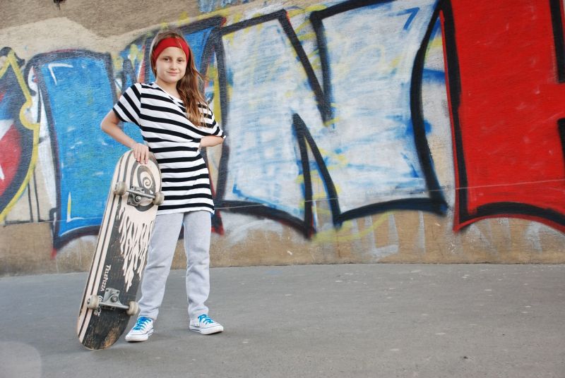 Girl in a striped shirt and red headband posing with a skateboard in front of some graffiti