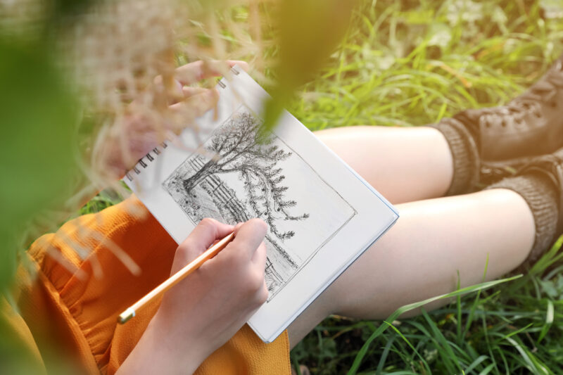 Young woman drawing with pencil in notepad while sitting on green grass, closeup