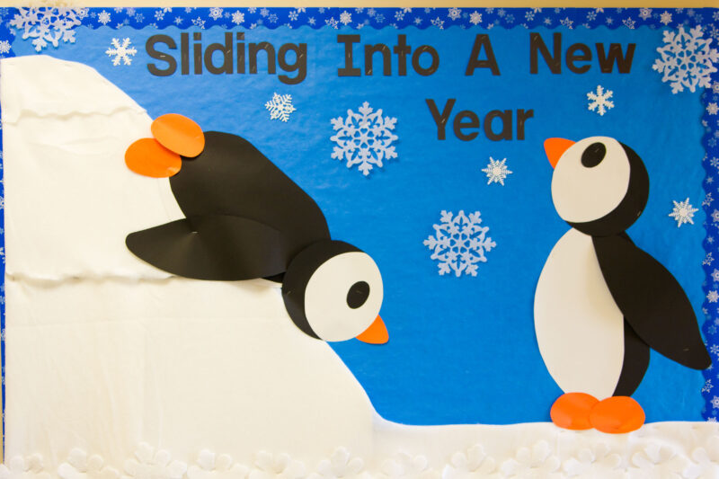A bulletin board shows two penguins, one is sliding down a snowy hill on its stomach. Text reads, "sliding into a new year." 