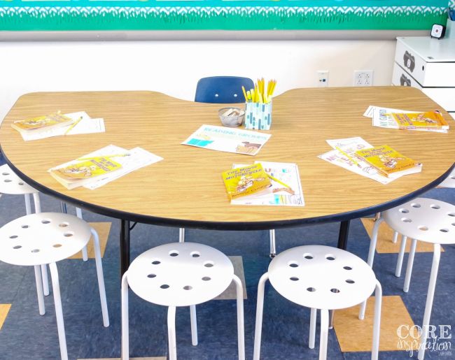 A small semicircular table with a teacher chair on the flat side and student stools around the outside edge, with books and other school supplies