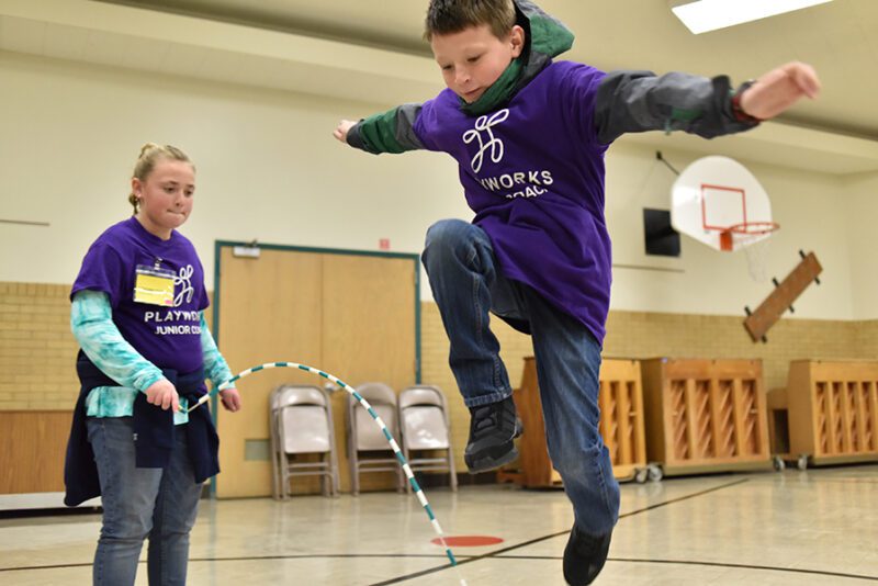 One student is shown shaking a jump rope while another student jumps over it. 