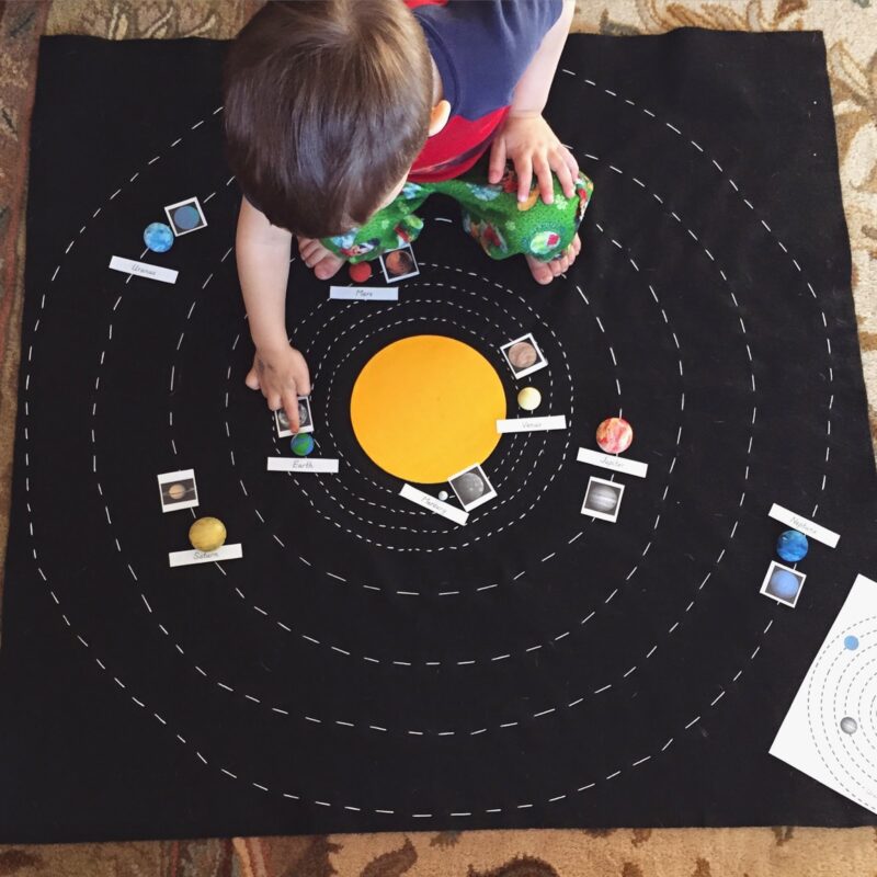 A little boy sits on a large black, felt map that has planets laid out on it and labeled (solar system projects)