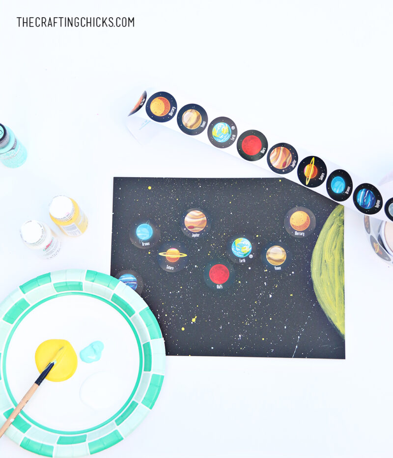 A plate has yellow paint on it with a paintbrush. A solar system scene has been painted and planet stickers have been stuck to it (solar system projects)