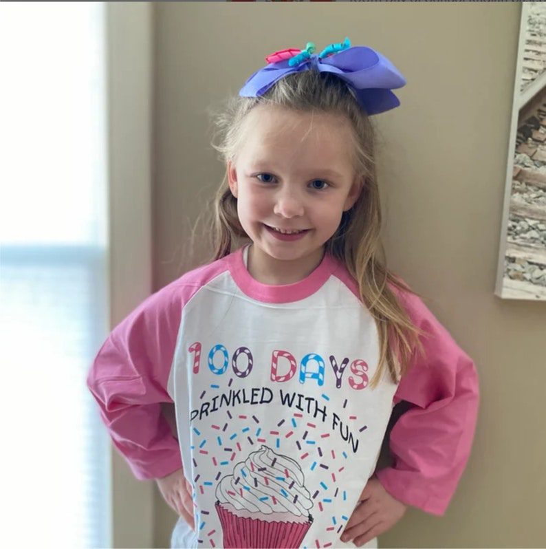 A little girl wears a shirt with pink sleeves that has a cupcake on it and says 100 Days Sprinkled with Fun. 
