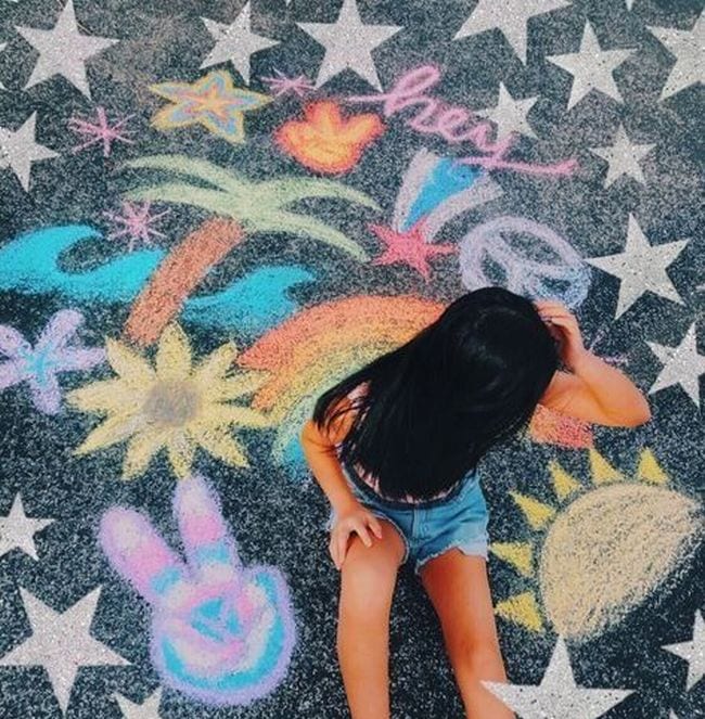Child sitting among sidewalk chalk drawings of palm tree, rainbow, sunshine, and more