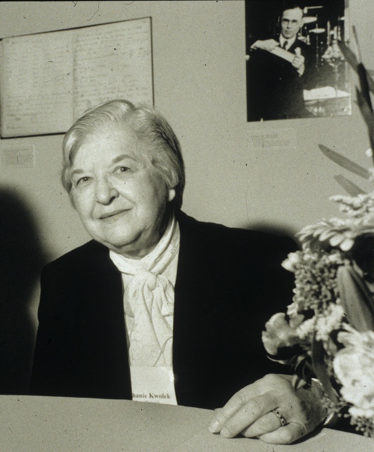 An older woman is seen nicely dressed from the waist up and seated at a desk.