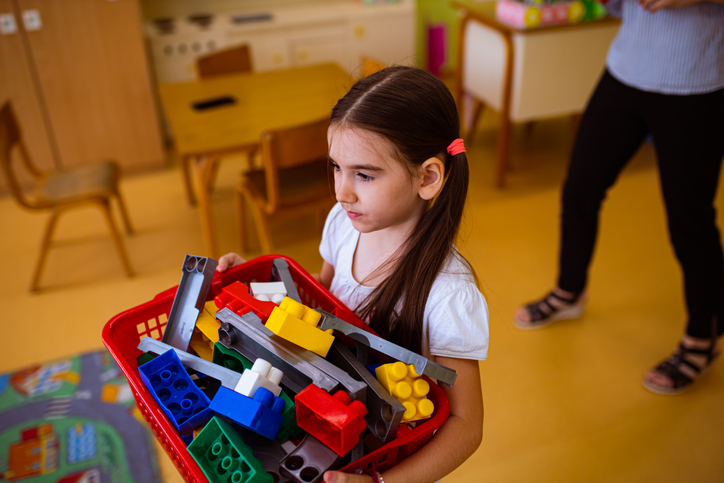 Young girl helps clean up bin of toys in classroom