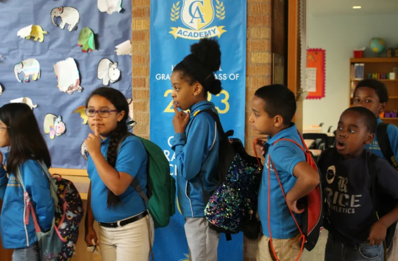 A diverse group of students wearing blue shirts and tan pants lined up to leave the classroom