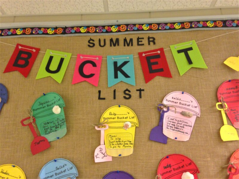 Text at the top of the bulletin board reads summer bucket list. It has different, brightly colored buckets and pails on it with students' writing assignments (summer bulletin boards ideas)