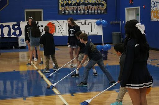 Pep rally activities include the one shown where students are sweeping a potato across a gym floor