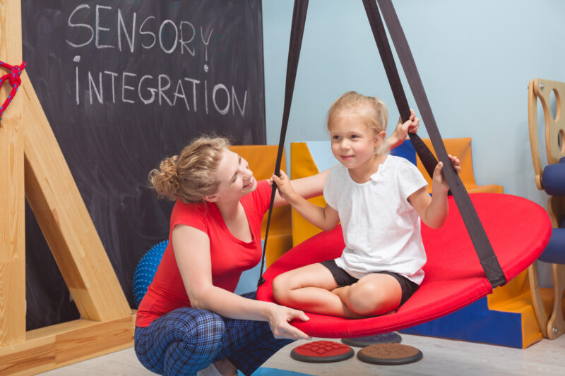 Shot of a smiling girl enjoying a sensory therapy on a swing while her physiotherapist assisting her