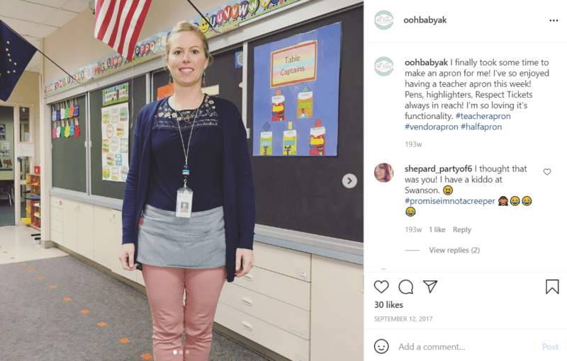 Teacher posing in her classroom in front of the chalkboard while wearing a plain, grey teacher apron