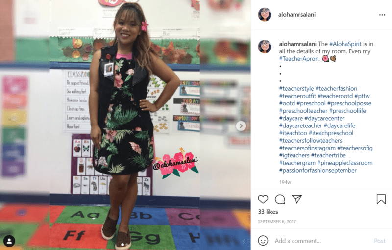 Teacher posing for a picture in her classroom while wearing a black apron with palm tree leaves and flowers on it embodying aloha spirit