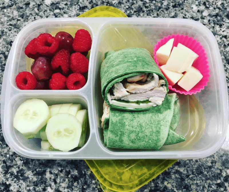 Divided container with a spinach chicken wrap, cheese cubes, cucumber slices, and raspberries