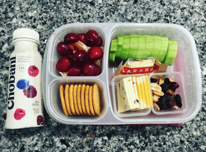 Drinkable Chobani Greek yogurt bottle next to a divided container filled with grapes, crackers, cheese, cucumber slices, and trail mix