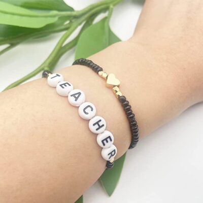A black bracelet with a gold heart and a black bracelet with beads that spell teacher are shown on a wrist.